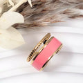 Bague dorée ajustable avec bande d’émail rose, acier inoxydable — présentation sur fond blanc et fleurs séchées.