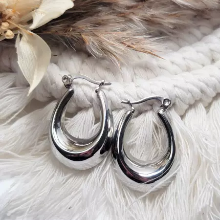Boucles d'oreilles créoles en acier inoxydable argenté sur un fond tissé avec des détails naturels.