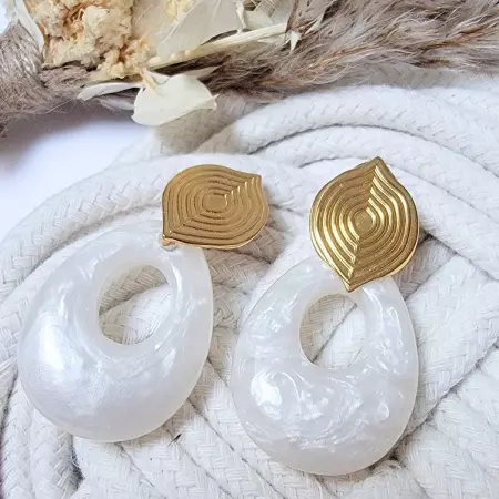 Boucles d’oreilles pendantes en résine nacrée et acier inoxydable doré, au design bohème chic, posées sur un fond tissé avec des fleurs séchées.