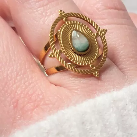 Bague en acier inoxydable doré avec une pierre naturelle amazonite en forme de goutte, posée sur un support blanc avec un fond décoratif de fleurs séchées et d’herbes pampas.
