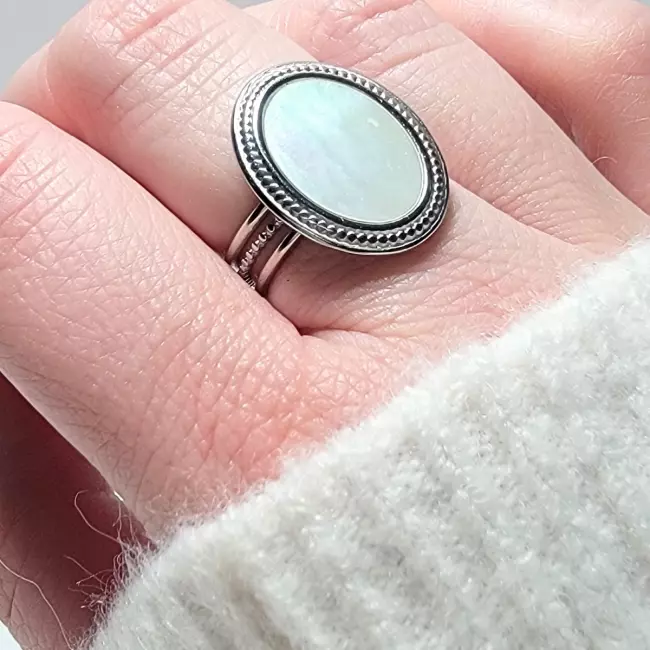 Bague en acier inoxydable argenté avec une pierre ovale en nacre naturelle, posée sur un fond blanc avec des fleurs séchées beiges